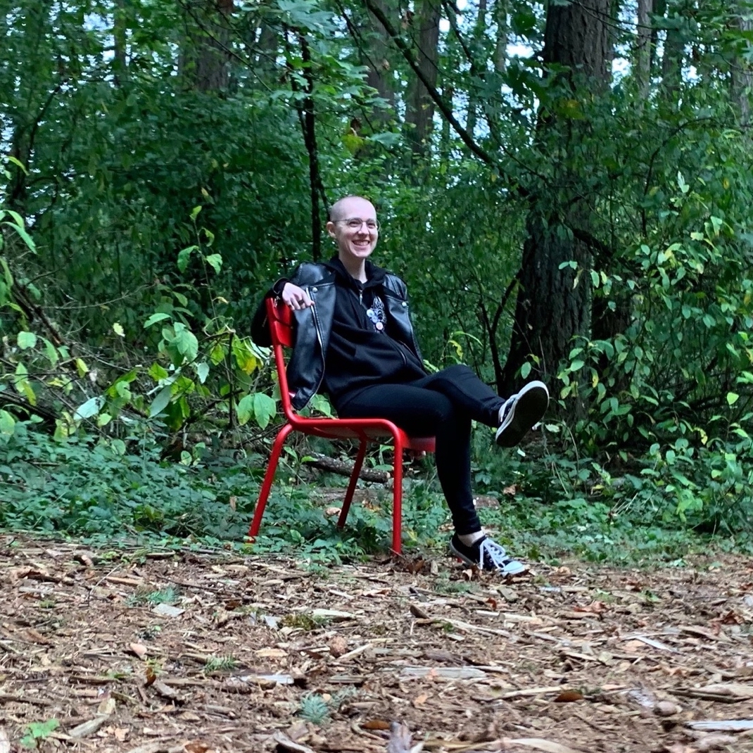 person with buzzed hair and glasses wearing a pleather jacket, black jeans, and black sneakers, sitting on a red chair in a clearing in the woods