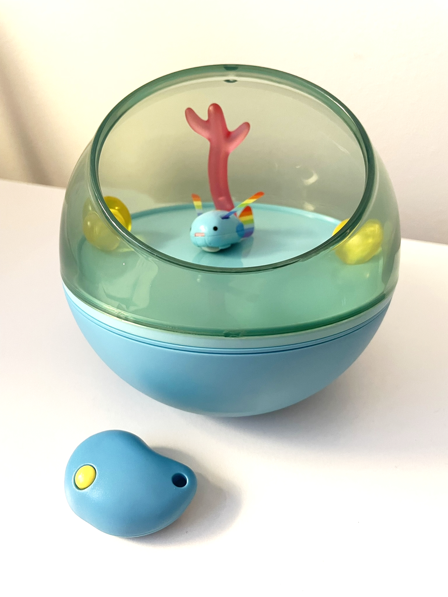 A blue orb, the top half transparent and with a large opening through which you can see several clear plastic coral pieces, and Mutsu themself - a cute fish-bunny hybrid with a blue body, rainbow ears and tail, and a yellow star on their forehead. To the side is the remote, made of blue plastic in the shape of a bend teardrop, with a single yellow button.